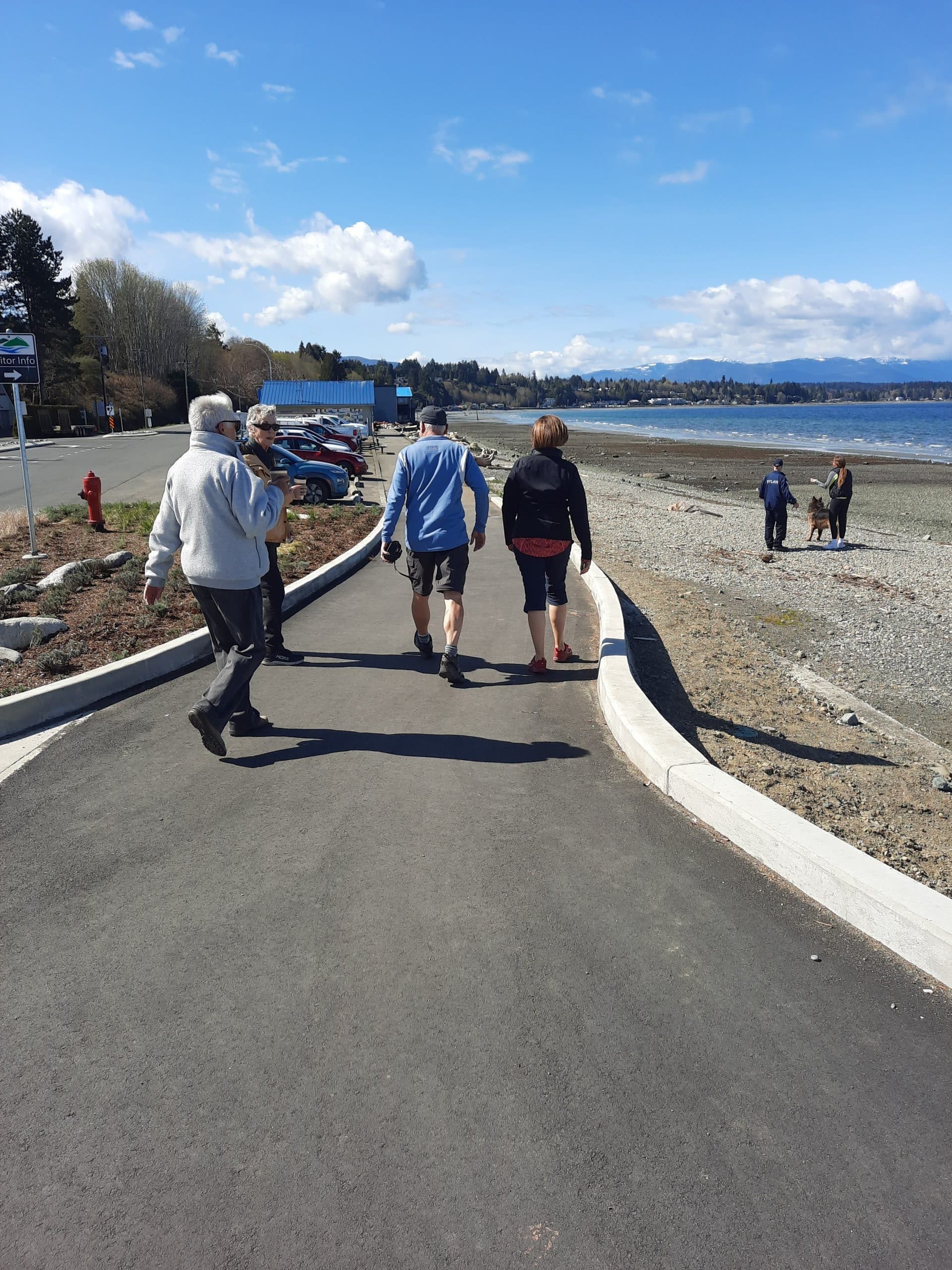 People walk on a cement trail beside the beach, with sand on one side and others enjoying the beach, under clear blue skies.