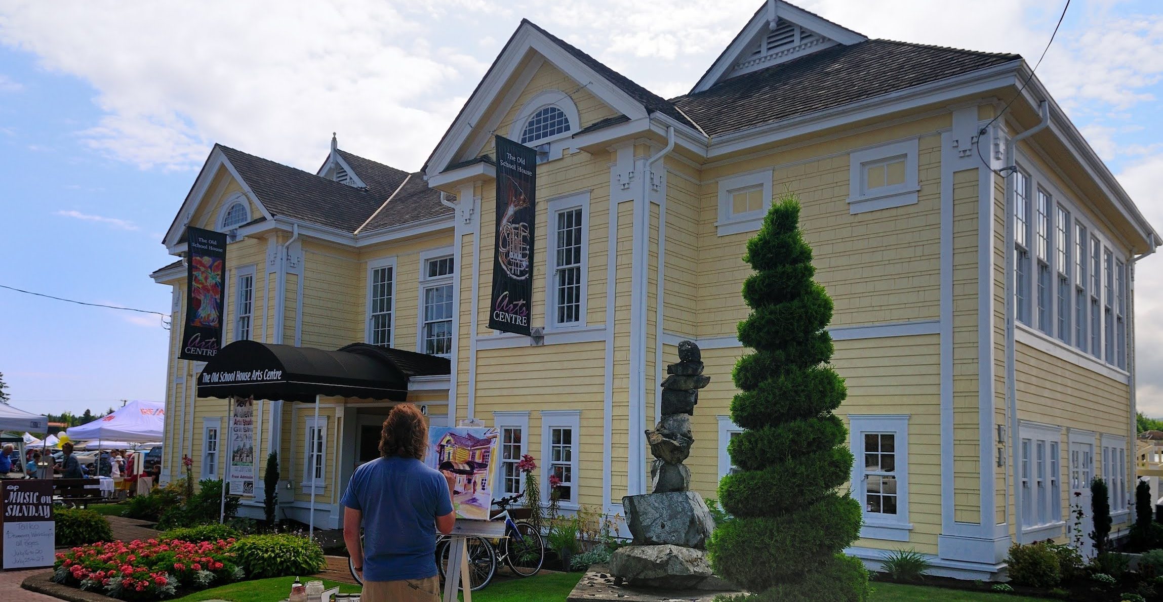 The Old School House Arts Centre Heritage Building, a yellow building with white trim, surrounded by trees and greenery. An artist is painting the building on an easel in front.