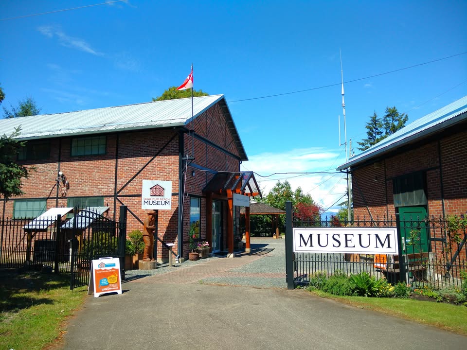 The entrance to the Qualicum Beach Historical and Museum Society, featuring an older brick building on either sides of the road entrance, and a gate sign that reads 