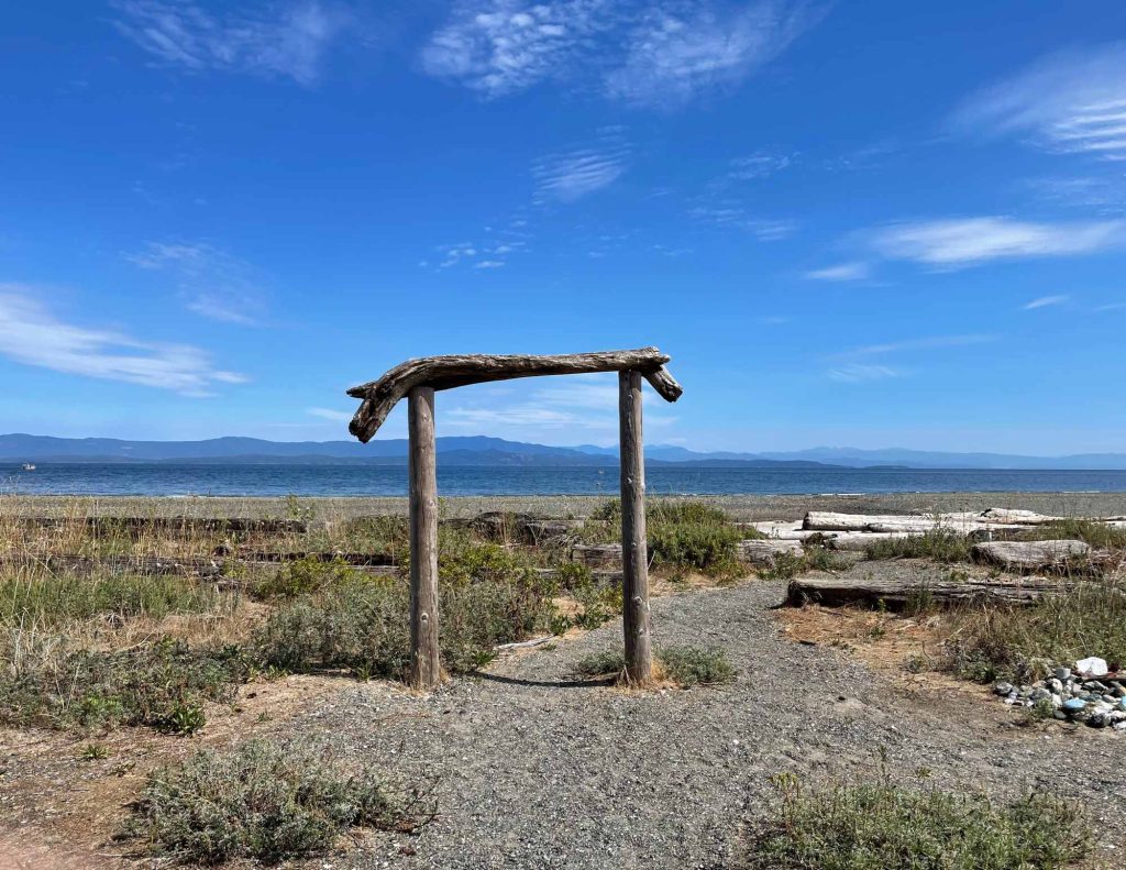 A park trail leading to the beach, with a wooden arch. There are clear blue skies, with mountains visible in the background.
