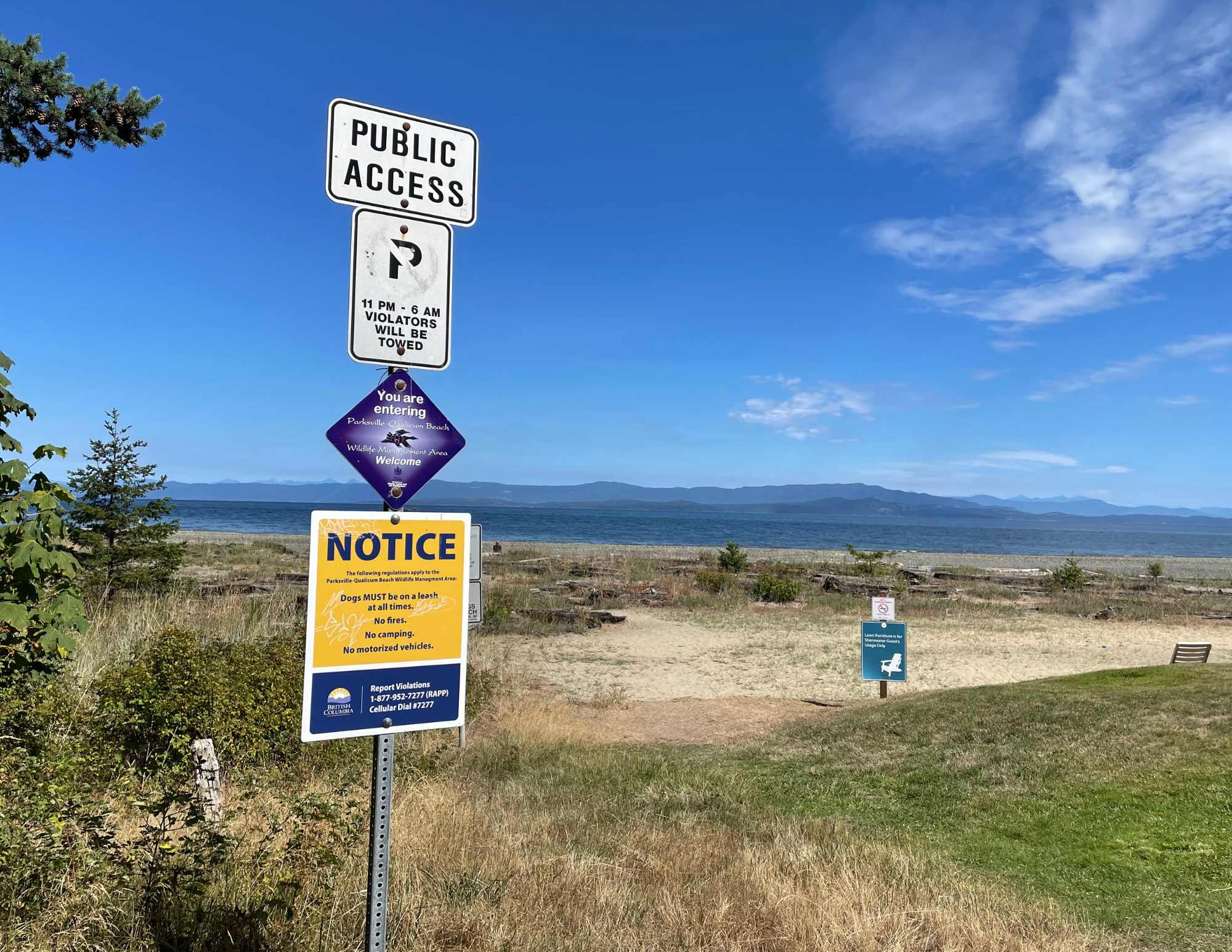 A grassy area leading to a sandy beach, with water and mountains in the background. A parking sign pole is visible in the foreground.