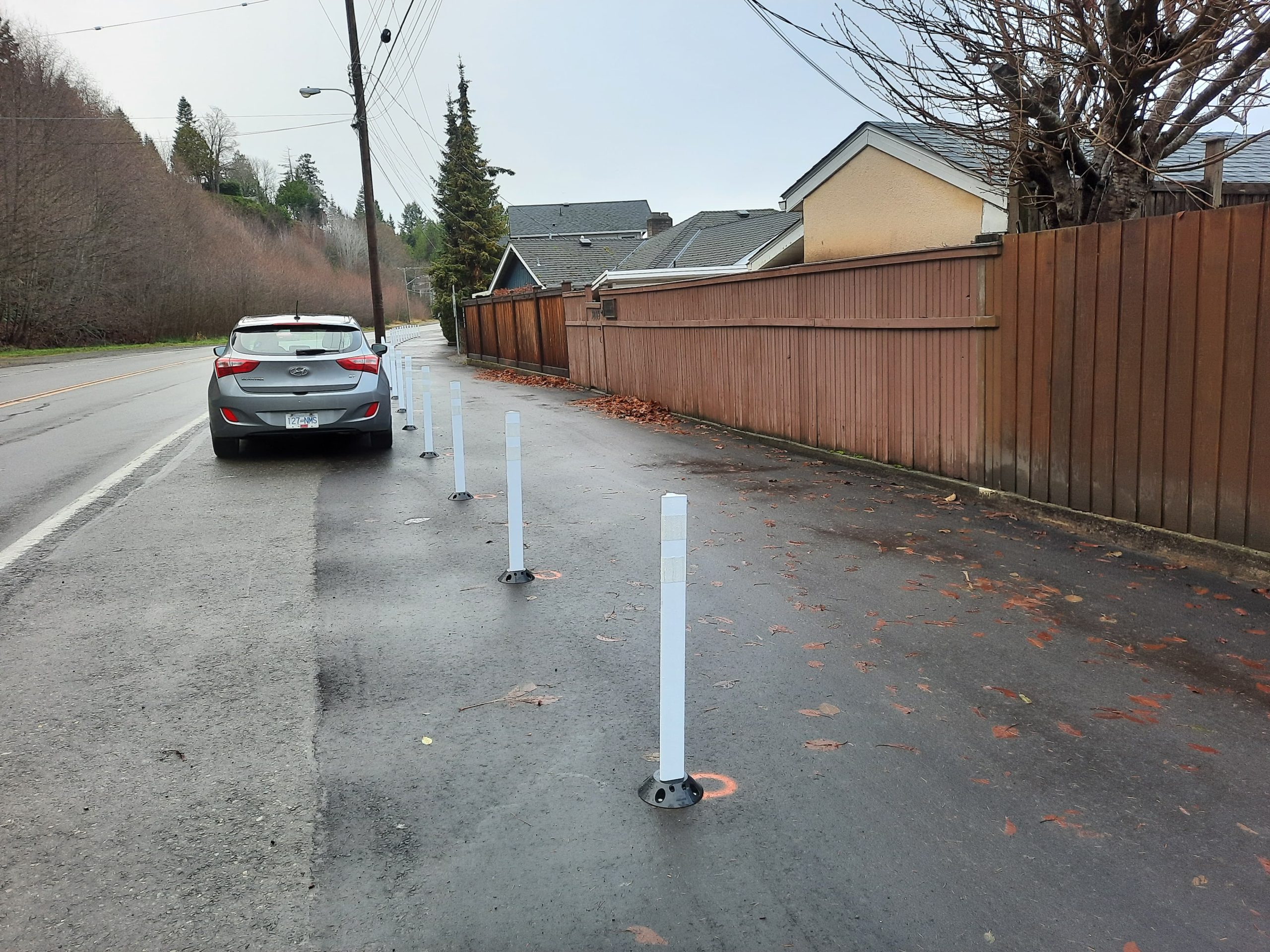 A walkway under construction, with a road on the left blocked by poles to prevent parking, and a fence on the right side of the path.