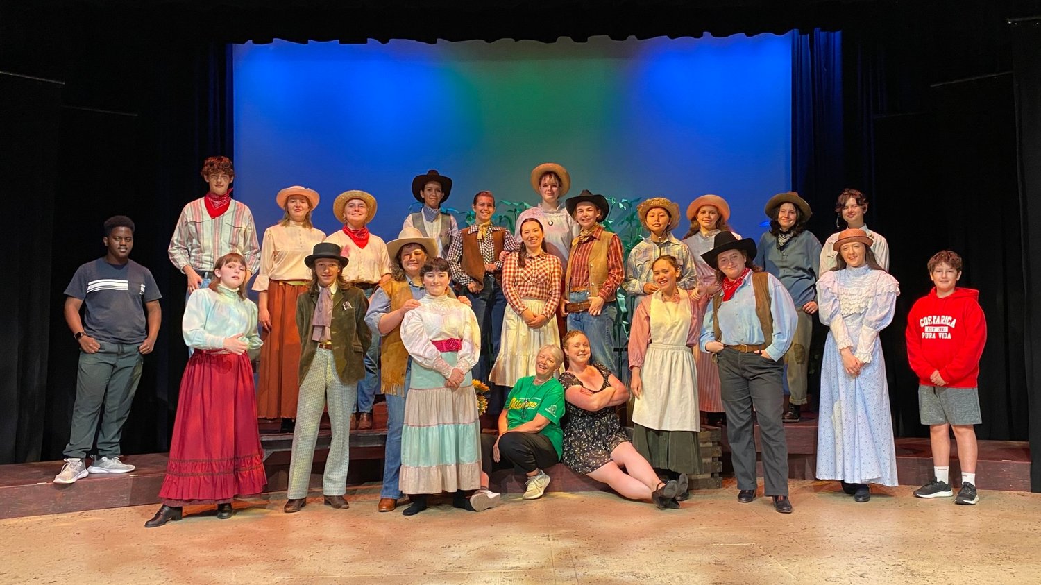 A group of people in costumes stands on stage, smiling after finishing a play.