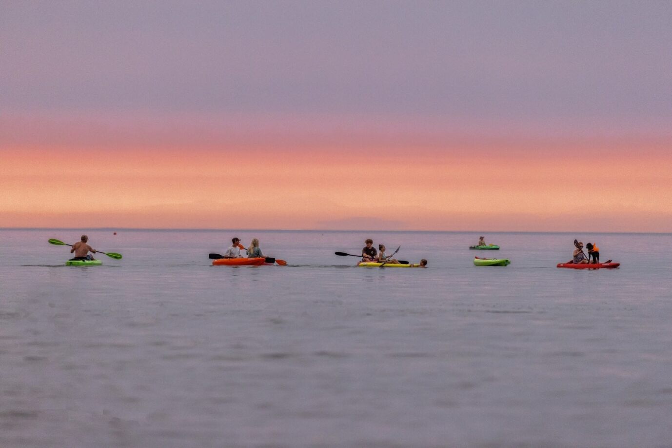 A group of kayakers in the water, with the sun setting behind them.