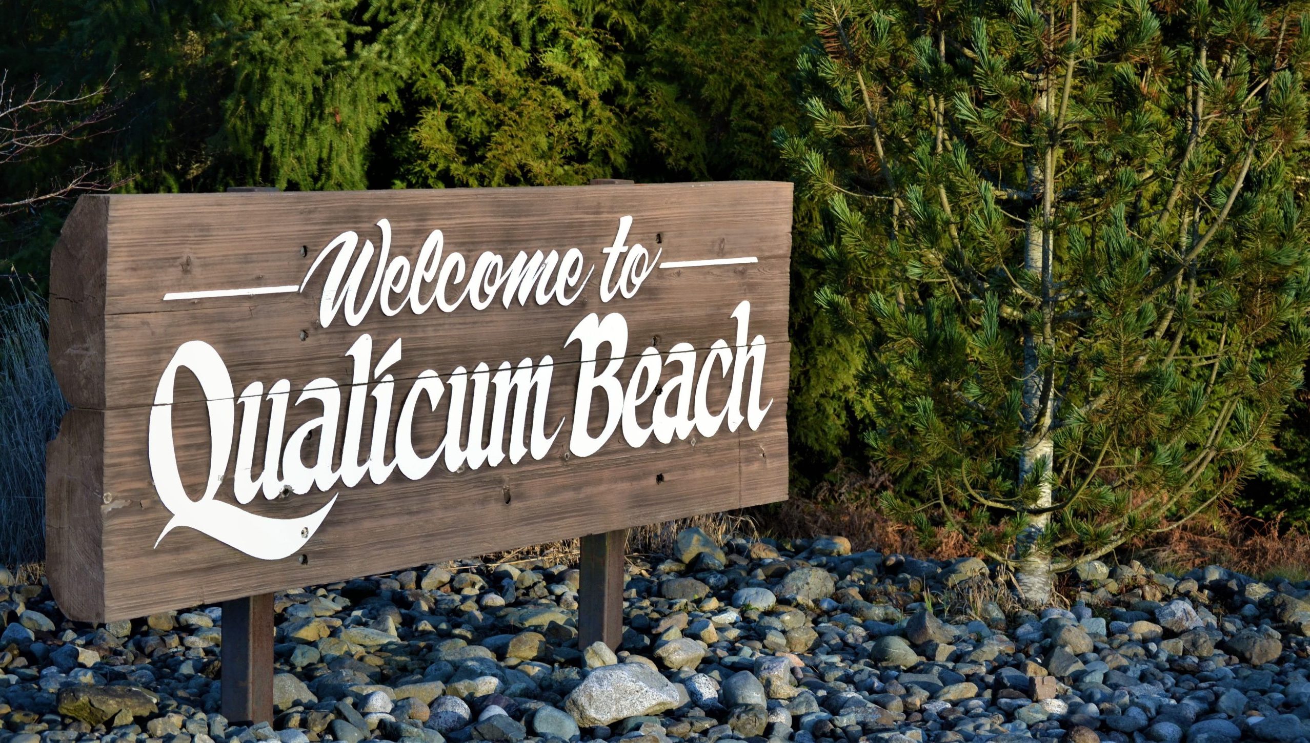 A dark brown wooden sign reads 'Welcome to Qualicum Beach' in white font. The sign is in a rocky ground and trees are around it.