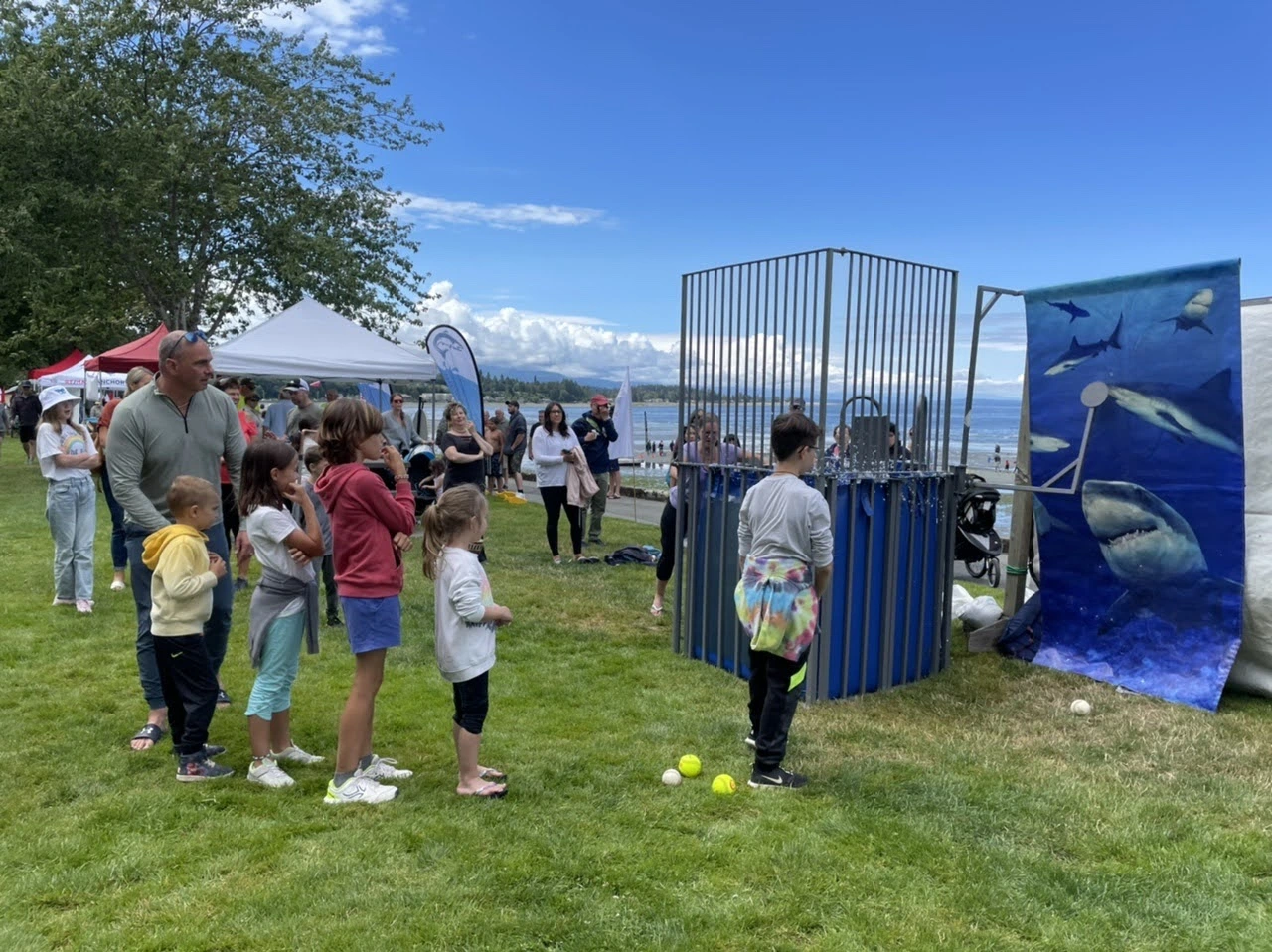 Children lined up in front of a shark themed dunk tank on a grassy lawn by the beach,