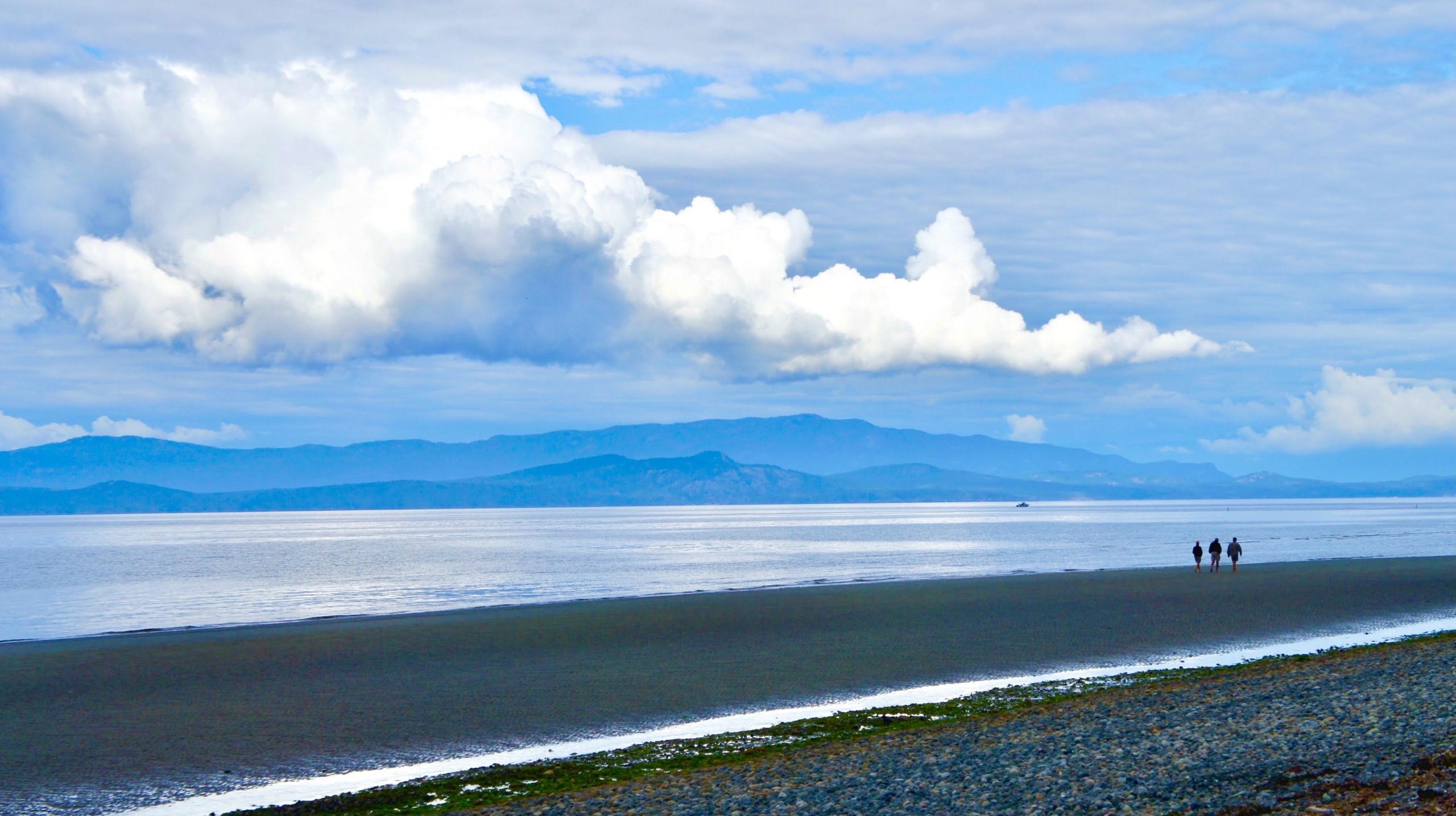 Three people are in the distance of an otherwise empty beach. The water is calm, there are mountains in the background, and the sky is filled with fluffy white clouds.