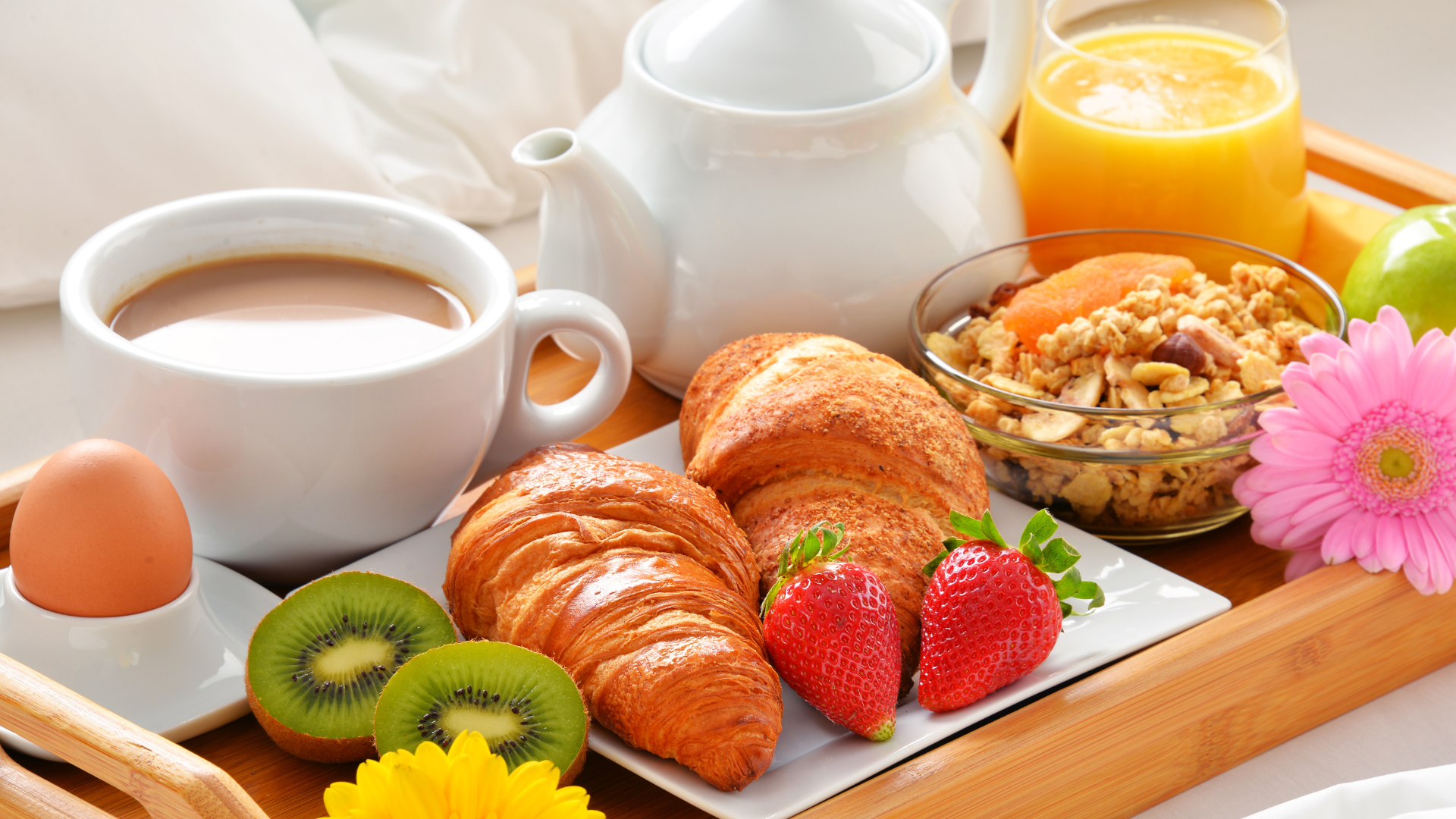 A breakfast tray is laid on top of white bed. The tray includes croissants, granola, fruit, an egg, coffee, a teapot and juice. It is decorated with flowers.