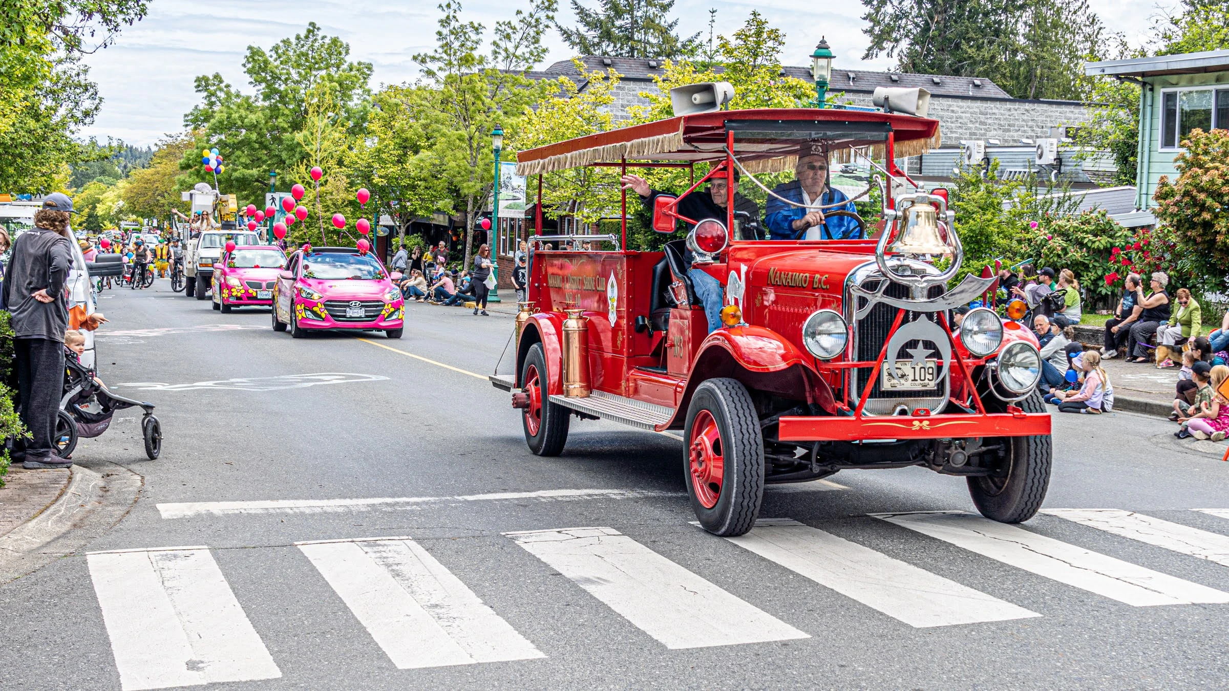 Red vintage vehicle leads a parade of pink cars for Family Day, decorated with balloons, down a street as parents and children watch.