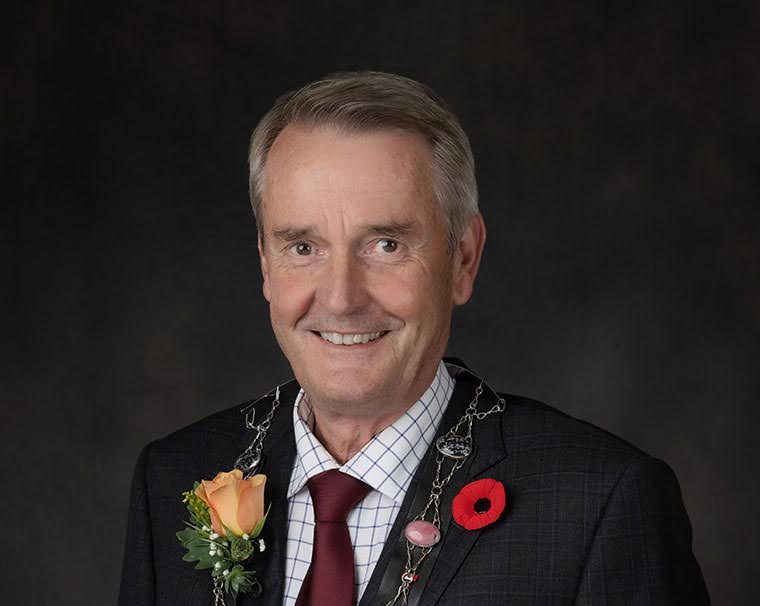 Mayor Teunis Westbroek against a dark grey background. Mayor Westbroek has short grey hair and is wearing a dark suit, checkered shirt, and burgundy tie. There is a red poppy pin on his left lapel, and a light orange rose on his right lapel. 