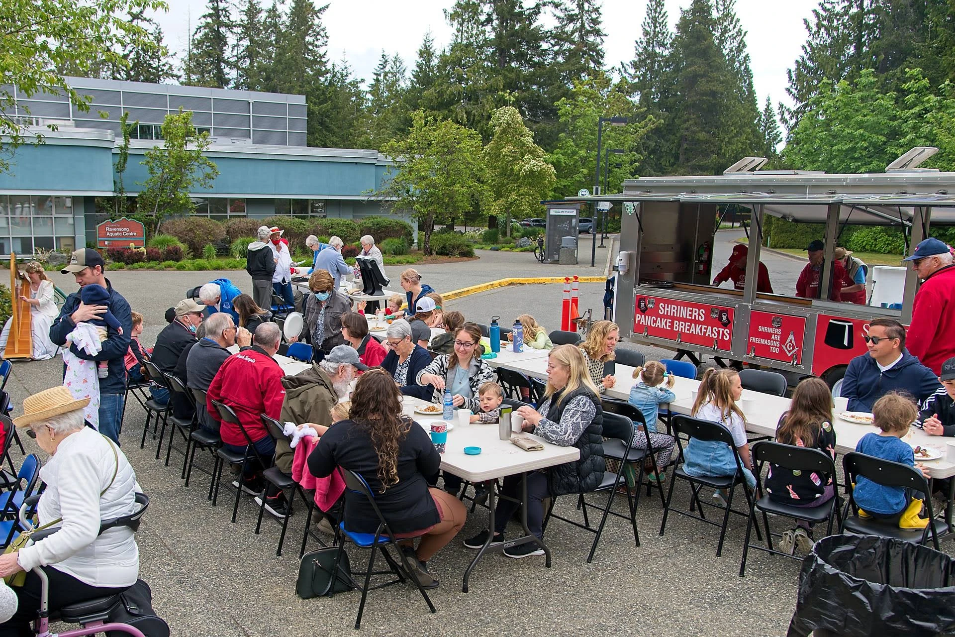 A large group of people enjoys a pancake breakfast event in a parking lot.