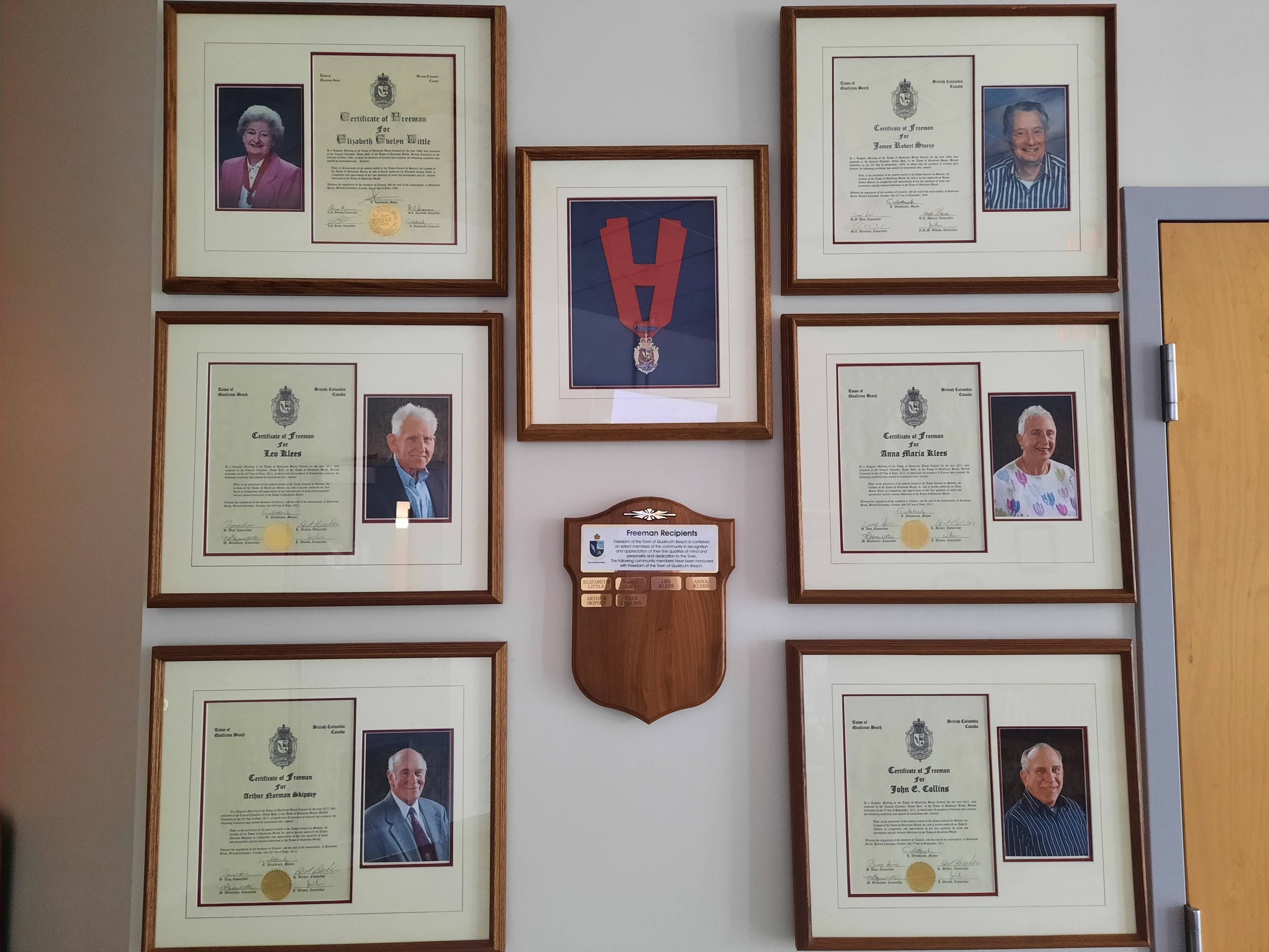 Six certificates, one medal, and a plaque hand on a white wall.