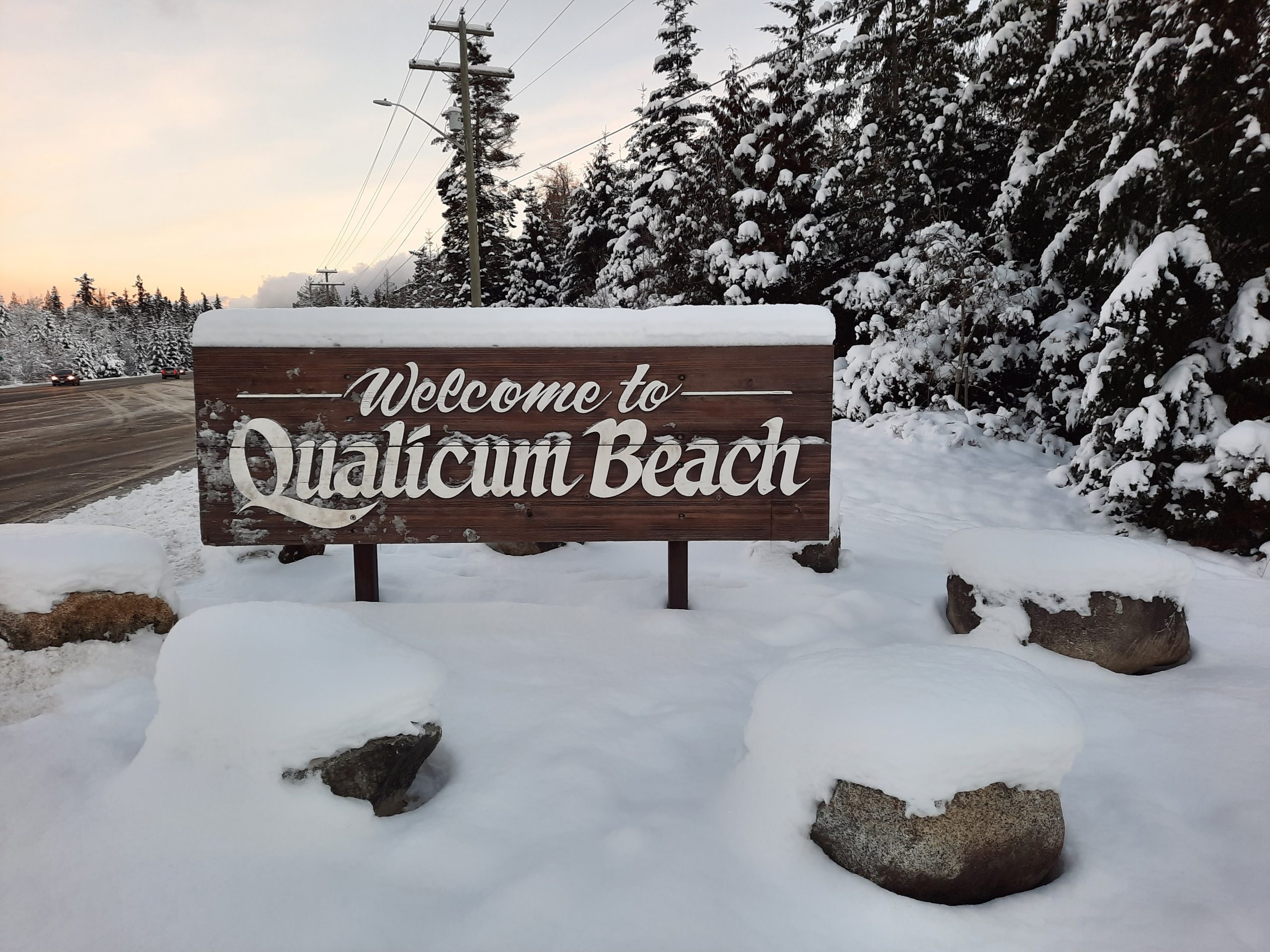 A dark brown wooden sign reads 'Welcome to Qualicum Beach' in white font. It is covered in snow, as is the ground and trees around it.